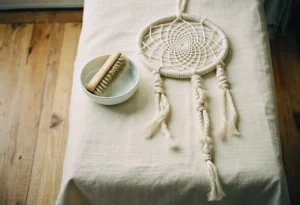 Attrape-rêves en macramé posé à plat sur un linge, à côté d'une brosse douce et d'un bol d'eau, prêt pour un dépoussiérage